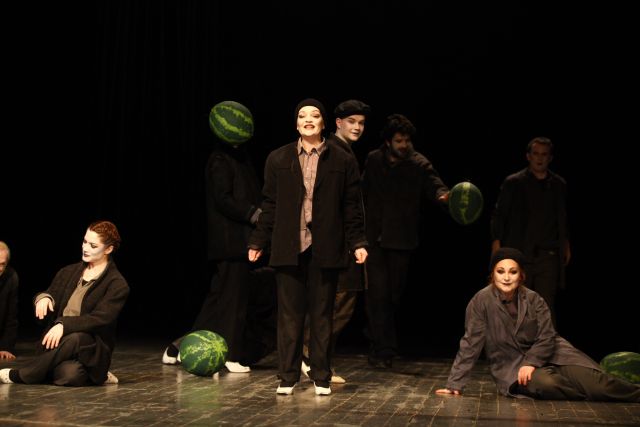 MOJA FABRICA I Schauspieler fotografiert von Muhamed Tunović Badi; 8 Schauspieler sind auf der Bühne zu erkennen. 3 von ihnen sitzen auf dem Boden, der Rest steht. Sie tragen alle dunkle Klamotten und ihre Gesichter sind weiß geschminkt mit schwarz gemalten Lippen und Augen. Einige von ihnen tragen auch eine schwarze Mütze. 2 Wassermelonen liegen auf dem Boden und 2 schweben in der Luft. 